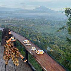 a woman sitting at a table with two plates of food on it, overlooking a valley