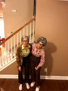 two older women standing next to each other in front of a stair case on a hard wood floor