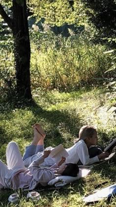 two people laying on the ground reading books in the shade under a shady tree and grass area