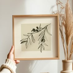 a person holding up a black and white print in front of a vase with dried plants