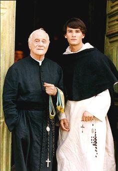 two men standing next to each other in front of a doorway with the priest wearing a black robe