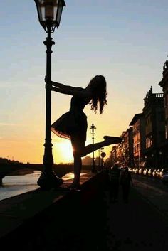 a woman is dancing on the street in front of a lamp post with a sunset behind her