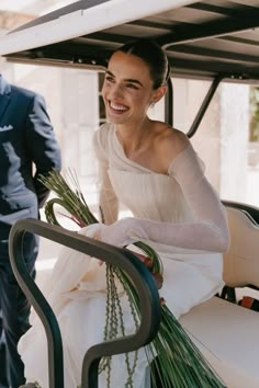 a woman in a white dress is smiling and holding flowers while standing next to a man