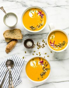 three bowls of carrot soup on a marble table with bread and spoons next to them