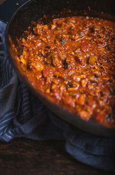 a pan filled with chili and meat on top of a wooden table next to a blue towel