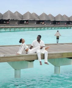 a man and woman sitting on a dock in front of some water with thatched huts