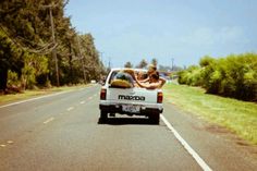 two people sitting in the back of a pickup truck driving down a road next to trees