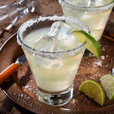 two margaritas on a tray with lime wedges and salt in the glass next to them