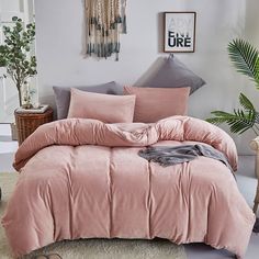 a bed with pink comforter and pillows in a room next to a plant on the floor