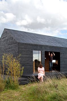 two people are standing on the porch of a small black cabin with open doors and windows