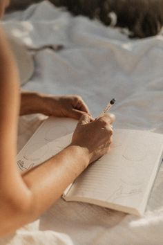 a person writing in a notebook with a cat laying on the bed behind them,