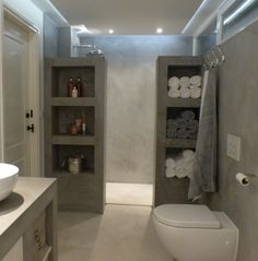 a white toilet sitting in a bathroom next to a sink under a mirror and shelves filled with towels