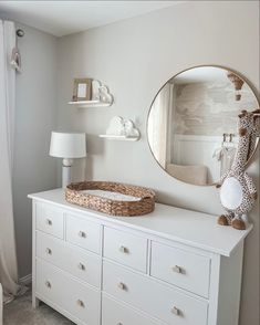 a white dresser with a basket on top and a round mirror above it in the corner