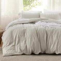 an unmade bed with white linens in front of a window and rug on the floor