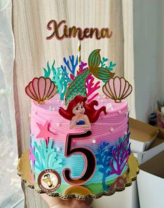 a hand holding a cake with the number five on it, decorated like a mermaid