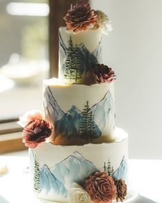 a three tiered cake decorated with flowers and pine trees on the top is sitting on a table