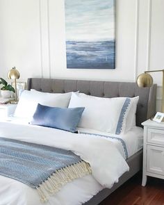 a bed with white sheets and blue pillows in a bedroom next to a painting on the wall