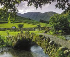 a stone bridge over a stream in the middle of a lush green valley with mountains in the background