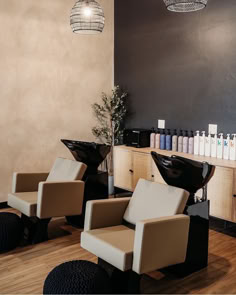 A salon space with two dark beige shampoo systems with black wash bowls and matching styling chairs standing in front of white wall-mounted styling stations with gold hardware and large mirrors. Salon Breakroom, Shampoo Station Ideas, Salon Seating Area, Salon Station Ideas, Salon Shampoo Area, Salon Shelves, Salon Vision Board, Rustic Salon, Hair Salon Stations