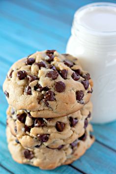 three chocolate chip cookies next to a glass of milk on a blue wooden surface with a jar of milk in the background