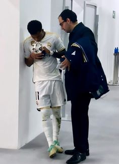 two men standing next to each other near a wall and looking at a cell phone