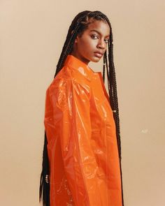 a woman with braids in an orange raincoat looking off to the side while standing against a beige background