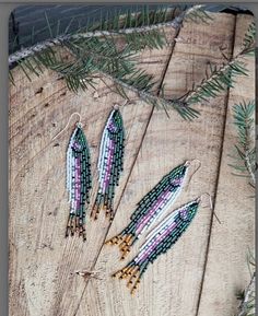 three beaded earrings sitting on top of a piece of wood next to pine needles