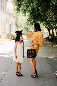 two girls in graduation gowns are standing on the sidewalk and pointing at each other