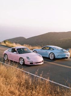 two pink sports cars driving down the road next to each other in front of some hills