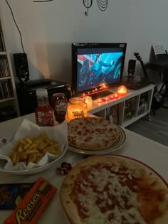 two pizzas and some fries on a table with a tv in the background,