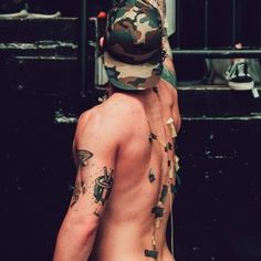 a shirtless man with tattoos on his chest wearing a camo hat and suspenders