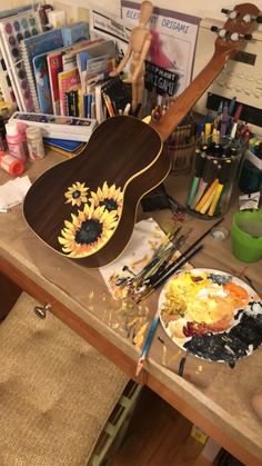 a guitar sitting on top of a wooden table covered in paint and brushes next to a painting palette