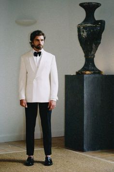 a man in a white tuxedo standing next to a black vase and floor lamp