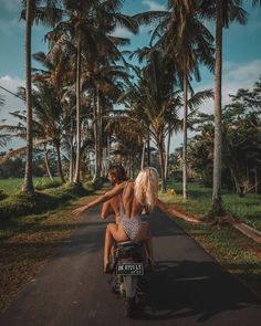 two people on a motor scooter riding down the road in front of palm trees