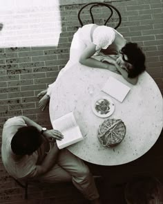 two people sitting at a table with books and papers on it, looking down at each other