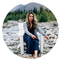 a woman sitting on a white chair in front of a mountain stream with trees and rocks