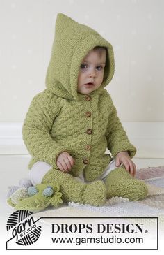 a baby wearing a green knitted hooded jacket and sitting on the floor with a stuffed animal