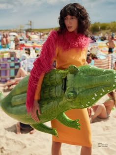 a woman in an orange dress is holding a fake alligator on the beach while people watch