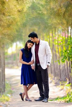 a man and woman standing next to each other on a dirt road surrounded by trees