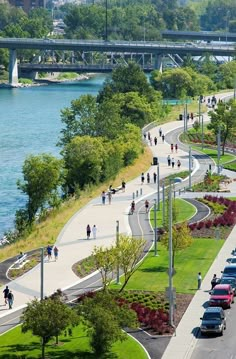 many people are walking on the sidewalk by the water and cars driving down the road