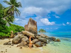 a woman standing on top of a sandy beach next to rocks and water with palm trees in the background