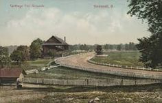 an old photo of a country road with a house on the other side and trees in the background