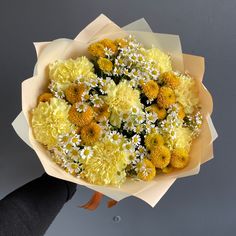 a bouquet of yellow and white flowers in someone's hand with gray wall behind