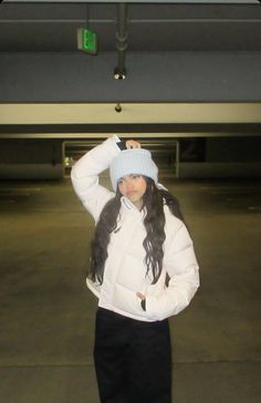 a woman standing in an empty parking lot wearing a white jacket and blue beanie