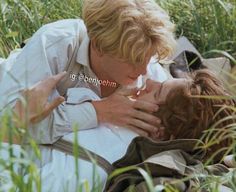 two people laying on the ground in tall grass, one is kissing the other's forehead