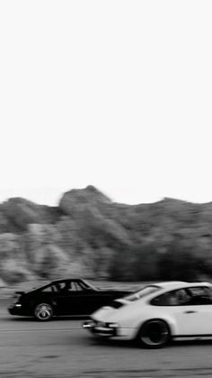 black and white photograph of two cars driving down the road with mountains in the background