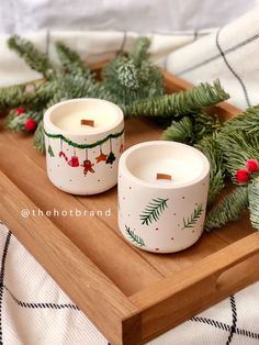 two white candles sitting on top of a wooden tray next to evergreen branches and pine cones