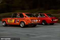 two orange cars driving down the road with trees in the background