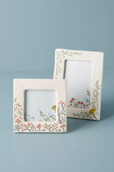 two white frames with flowers and birds painted on them, sitting side by side in front of a blue background