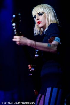 a woman with blonde hair and black glasses holding a guitar in her right hand while standing on stage